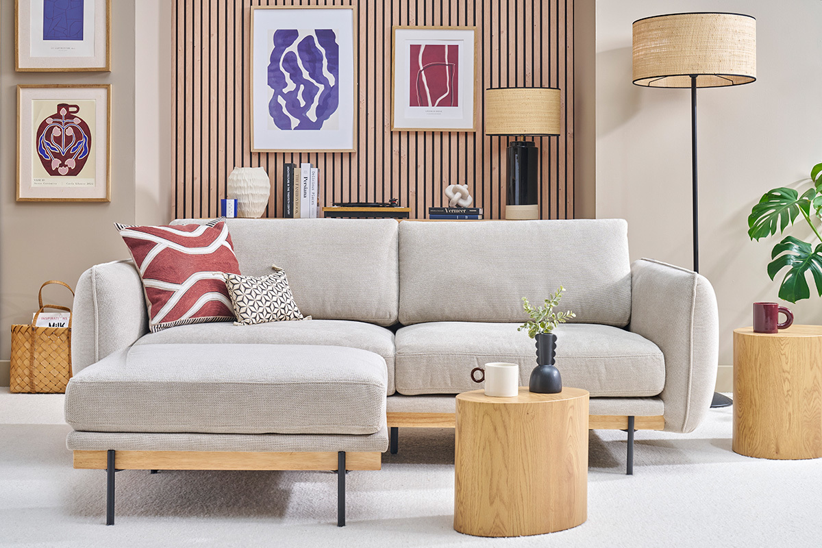 Canap beige 3 places avec repose-pieds, angle frontal, entour de tables en bois et dcor mural moderne.