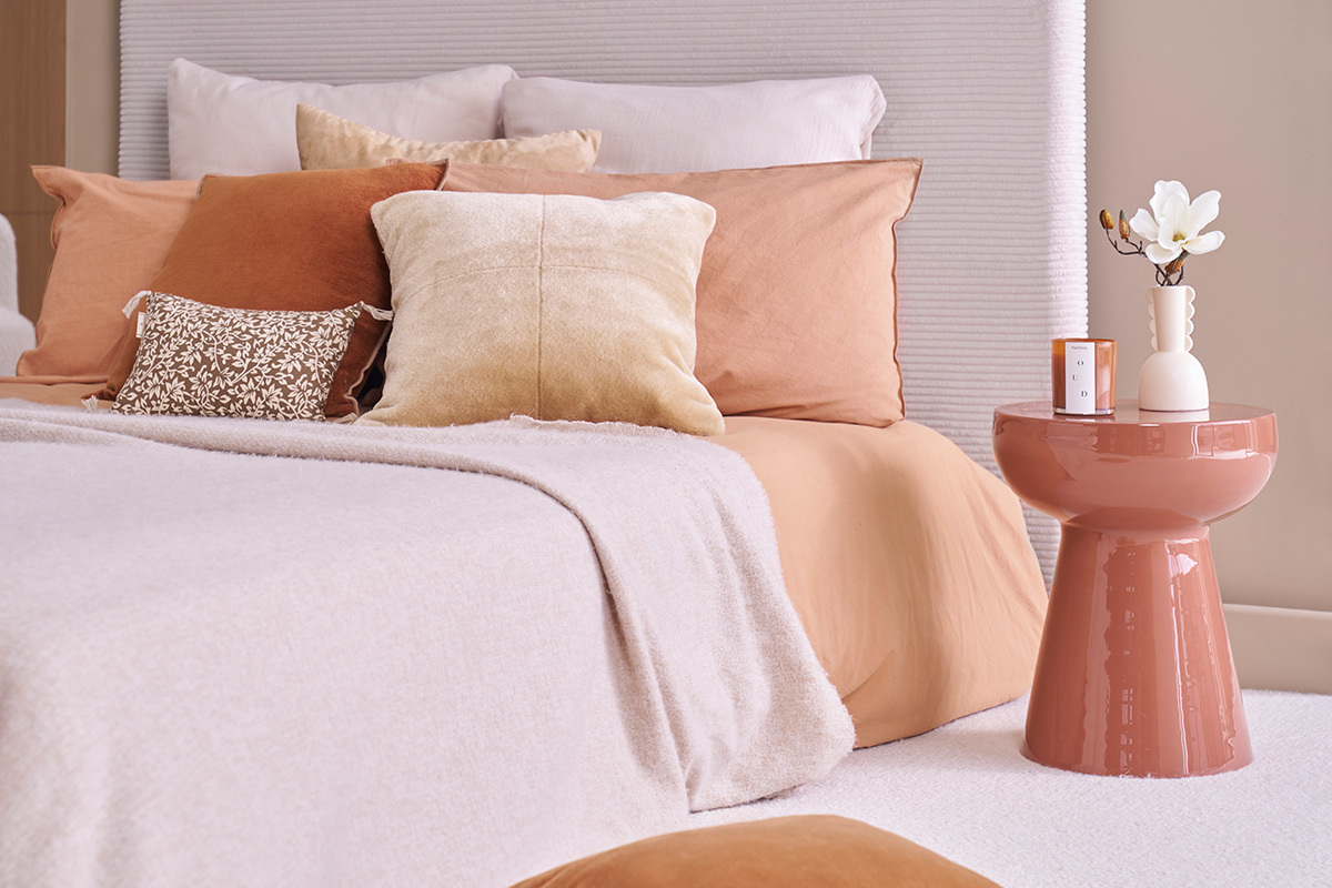 Table d'appoint ronde en m�tal �maill� terracotta � c�t� d'un lit avec des coussins beiges et terracotta.