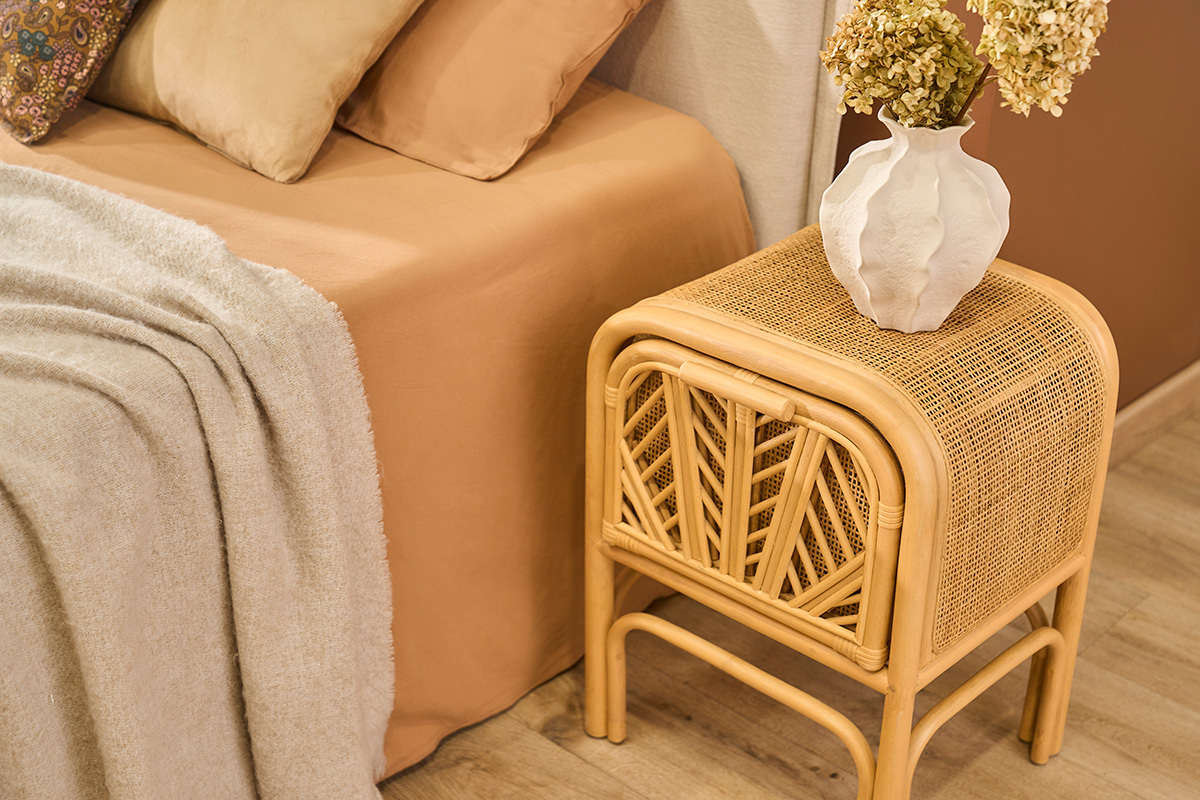 Table de chevet en rotin GALON, vue de c�t�, avec vase, � c�t� d'un lit beige.