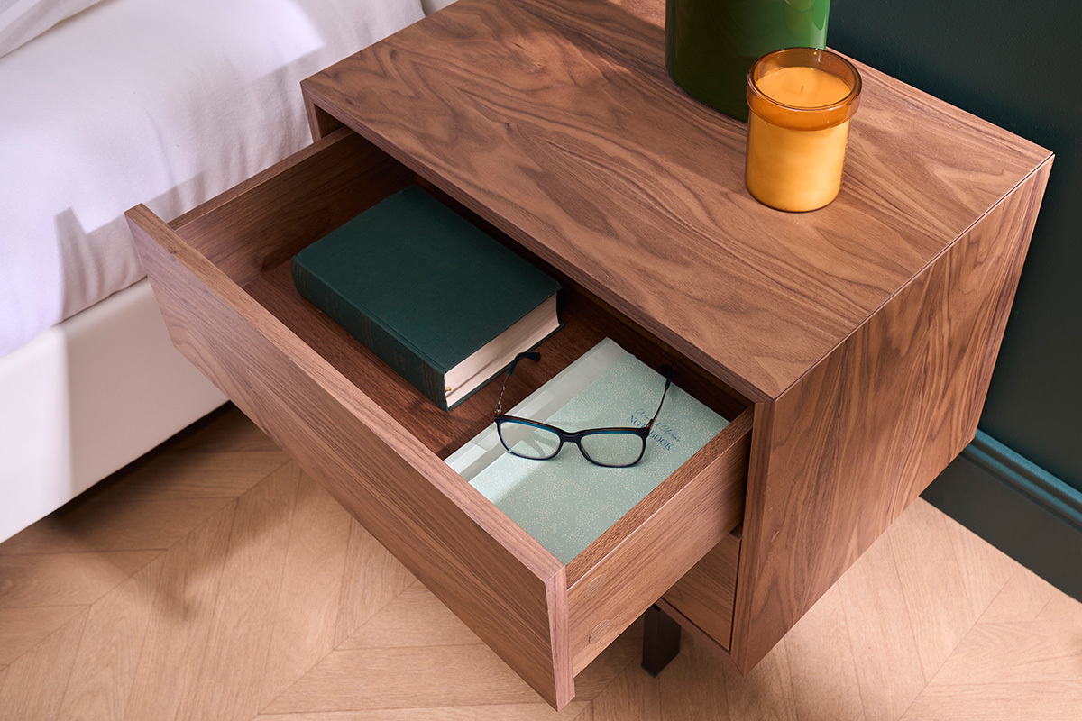 Table de chevet bois fonc avec tiroir ouvert contenant livre et lunettes.