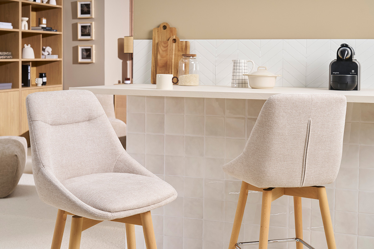 Deux tabourets de bar scandinaves en tissu beige et bois clair devant un comptoir de cuisine.